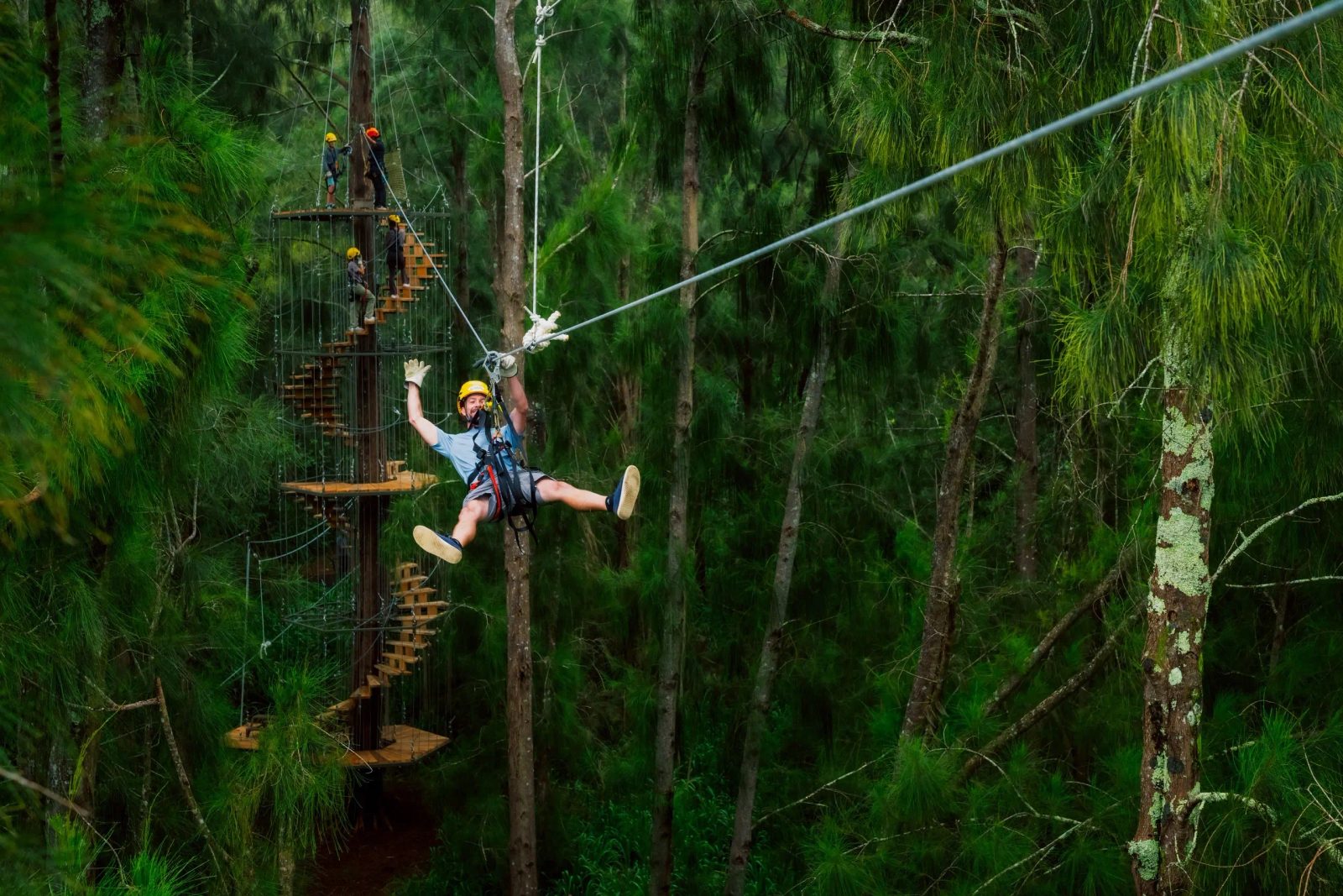 Guest zip-lining through a pine forest with arms raised in excitement, a multi-level treetop tower and spiral staircase visible behind him on the Kohala Zip & Dip.
