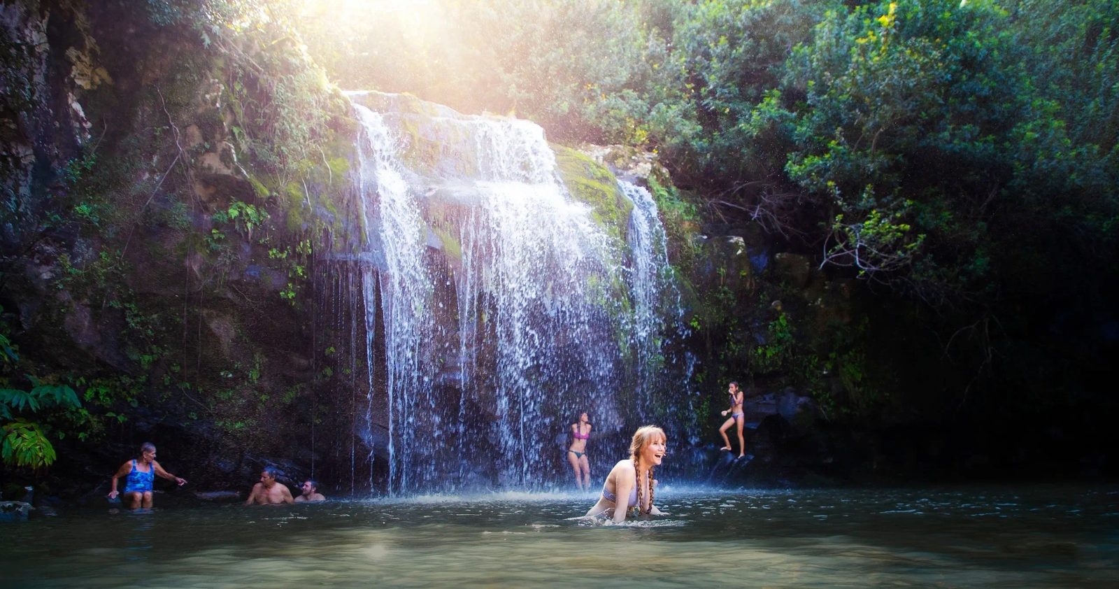 Guests swim and laugh in a jungle waterfall pool on the Kohala Zip & Dip, with a wide waterfall cascading down mossy cliffs behind them.