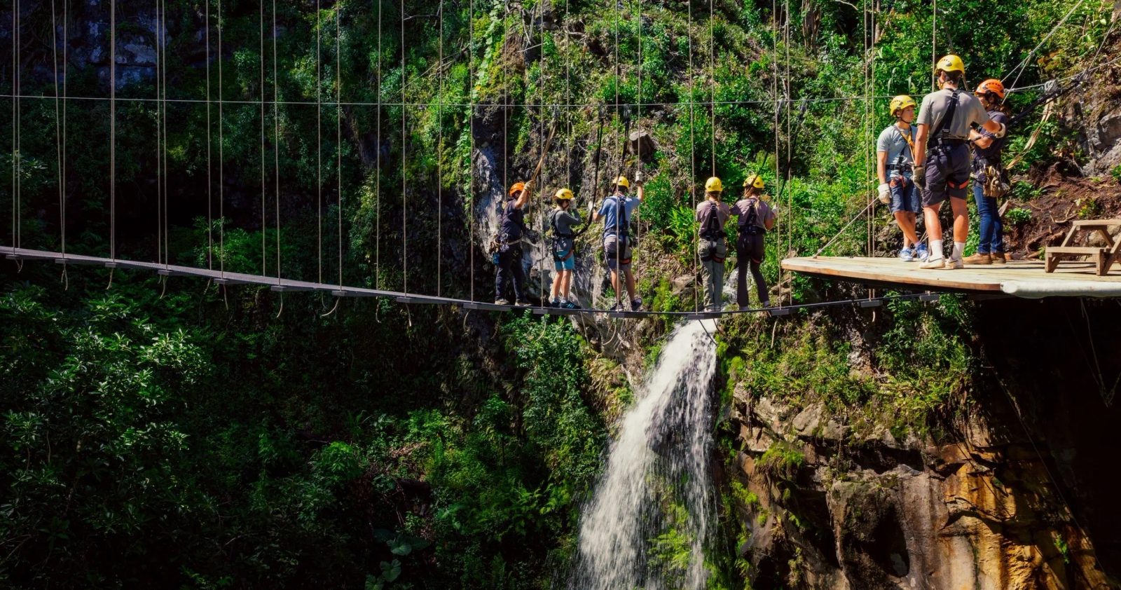 Helmeted guests cross a suspended sky bridge high above a waterfall and lush ravine on the Kohala Zip & Dip.