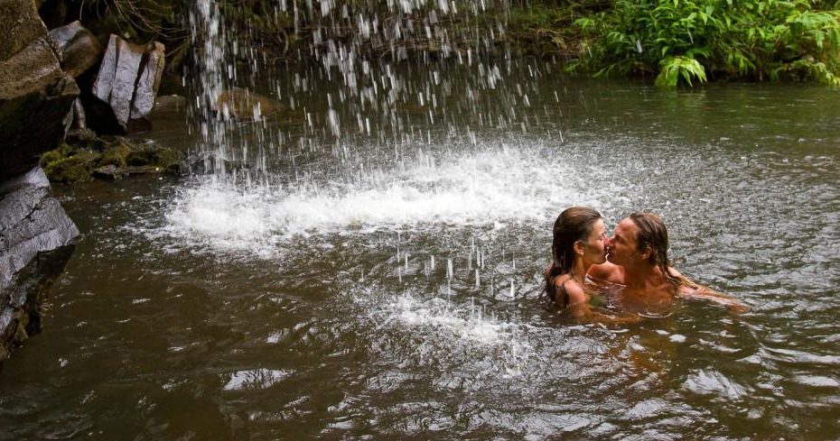 A couple embraces beneath a cascading waterfall in the swimming hole on the Kohala Zip & Dip.