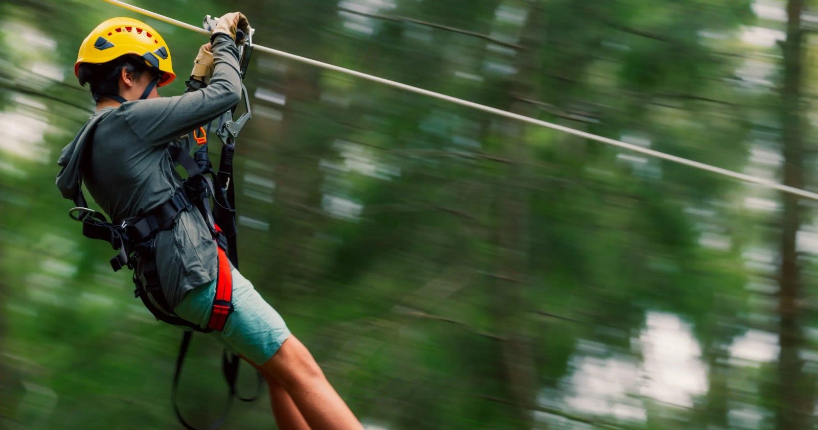 Close-up of a guest in a yellow helmet zip-lining at speed, forest blurred behind him.
