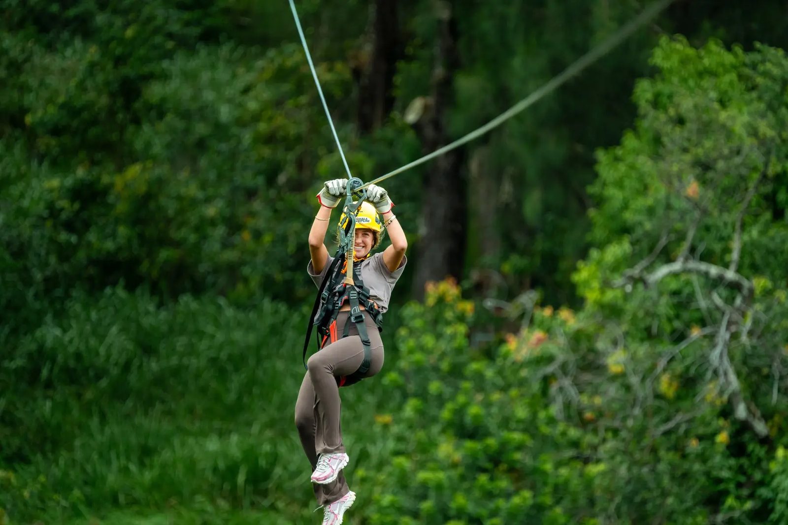 Guest in a yellow helmet zip-lining through a North Kohala forest, arms raised overhead gripping the trolley.