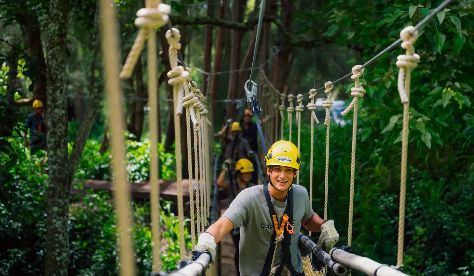 Smiling guest in a yellow helmet crossing a rope suspension bridge through the forest, other guests following behind.