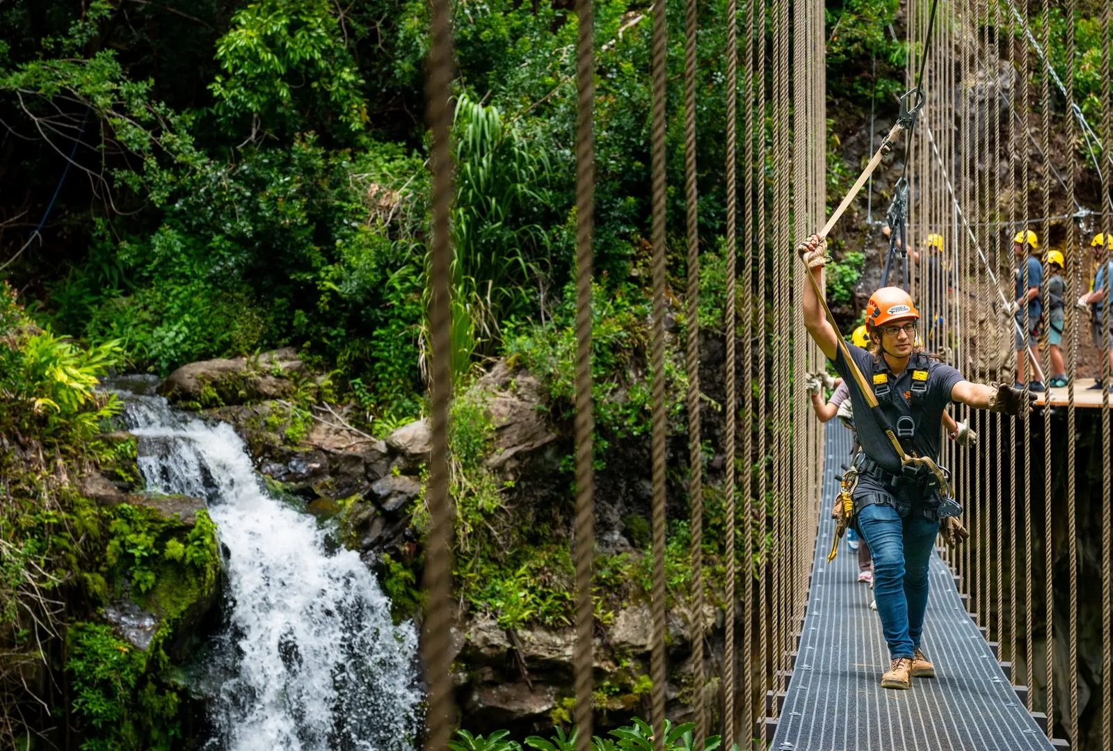 Guest in an orange helmet crossing a metal sky bridge beside a waterfall cascading down a moss-covered cliff.