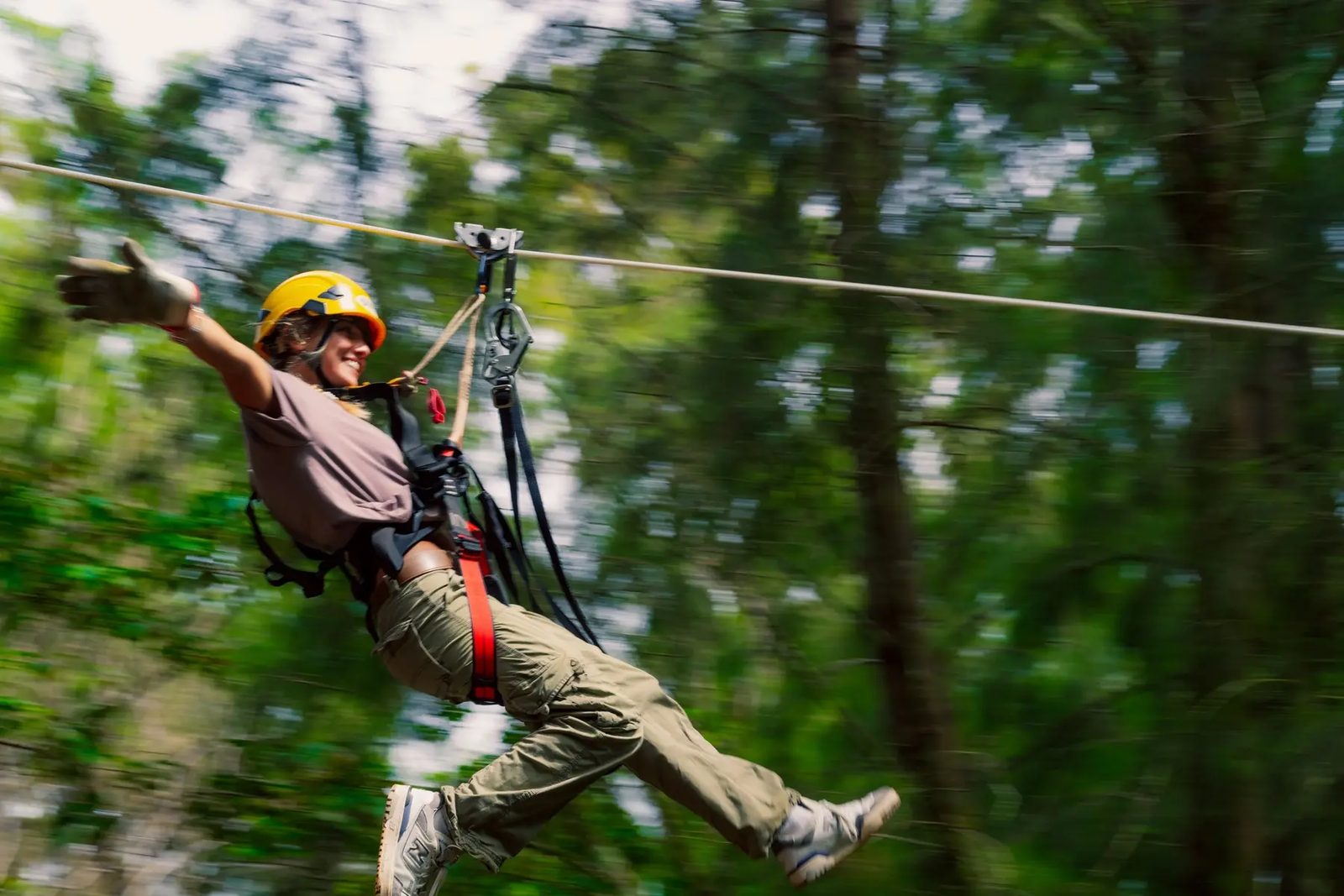 Guest in a yellow helmet zip-lining at speed with one arm raised, forest blurred by motion behind her.