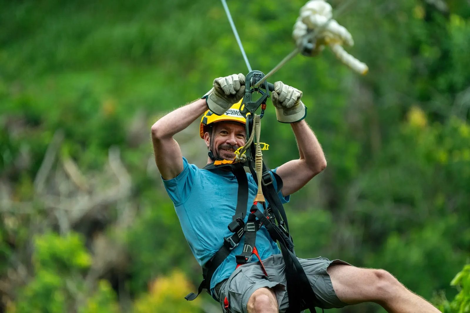Smiling guest holding the trolley overhead on a Kohala Zipline course cable, forest visible behind him.