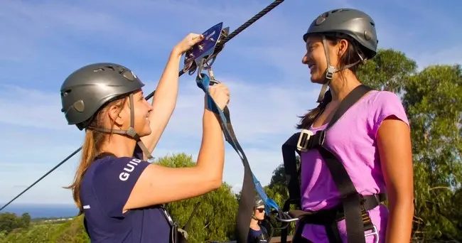 Guide clips a guest's harness onto the zipline cable before launch, with the Hāmākua coastline visible in the background.