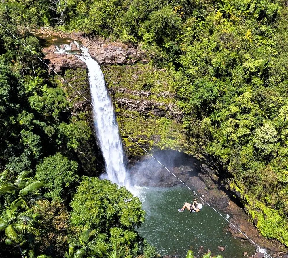 Aerial view of a guest zip-lining across a jungle pool beneath a tall waterfall on the Kolekole Waterfall Zipline Adventure.
