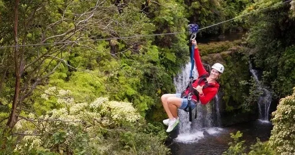 Guide zip-lining over a waterfall on Hawaii Island, arm raised with a thumbs up, cascading falls and lush jungle behind her.