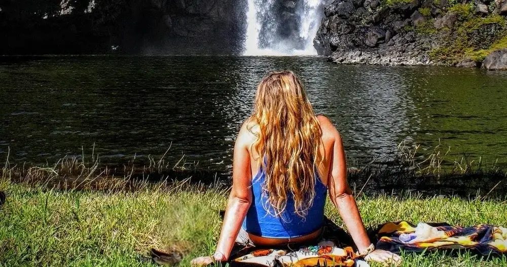Guest sitting on the grass at the edge of a private waterfall pool on Hawaii Island, waterfall cascading into the water ahead of her.