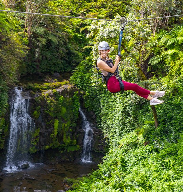 Smiling guest zip-lining above a double waterfall in a lush jungle ravine on Hawaii Island.