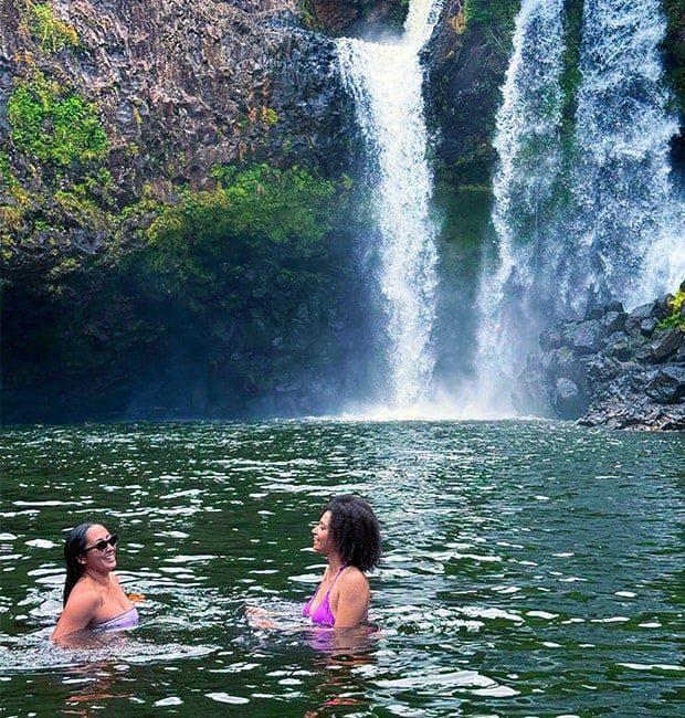 Two guests swimming and laughing together in a private waterfall pool on Hawaii Island.