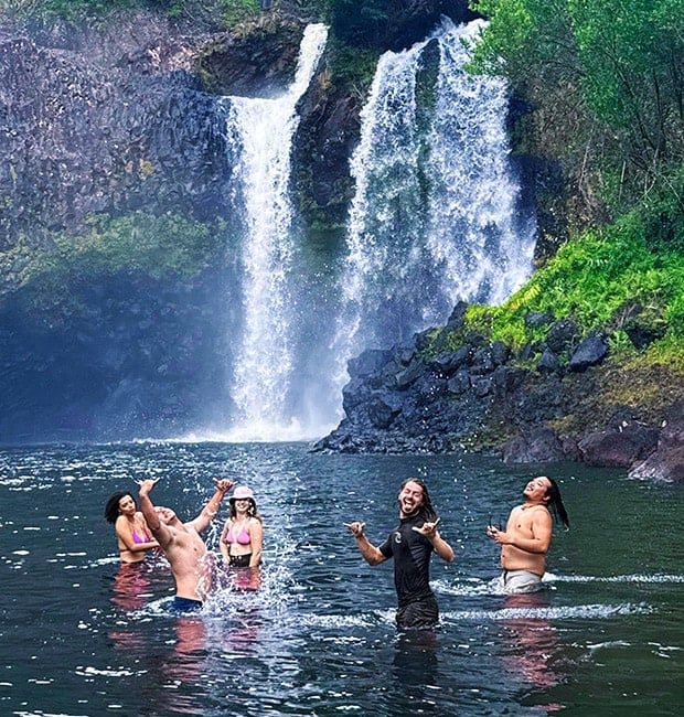 Group of guests celebrating and splashing in the pool beneath a double waterfall on Hawaii Island.