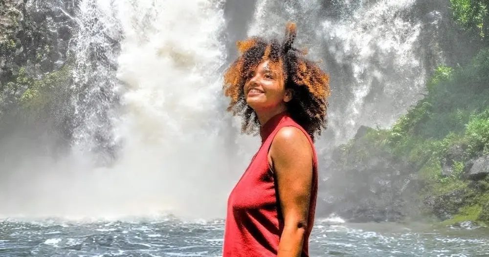 Woman stands beside a large tropical waterfall and dark freshwater pool on Hawaii Island, with mist rising from the falls behind her.