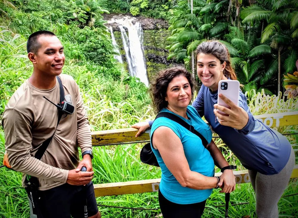 Guide and two guests smiling and taking a phone selfie in front of a cascading waterfall on Hawaii Island.