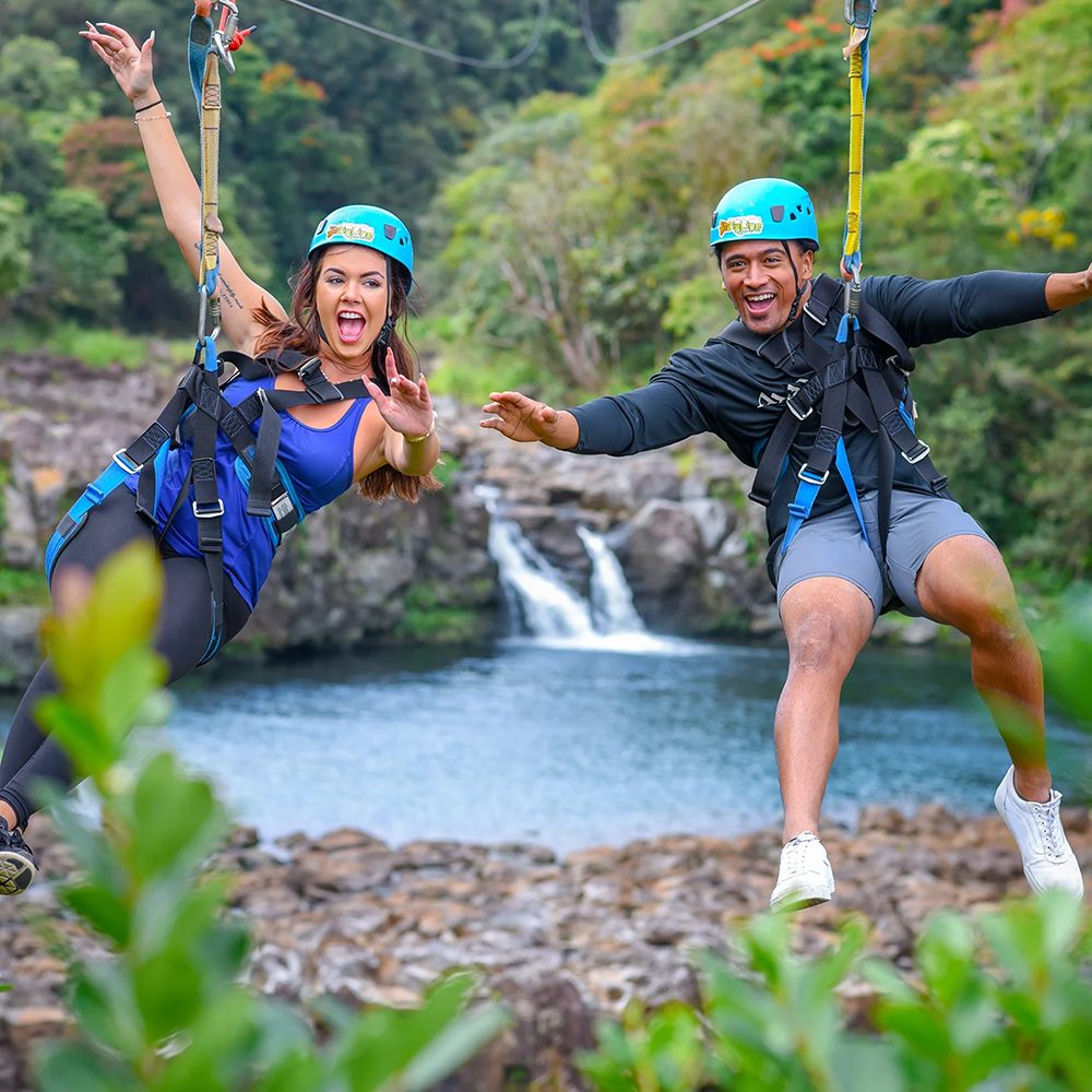 Two zipline guests laughing and reaching toward each other on side-by-side dual lines above a waterfall pool at Umauma.