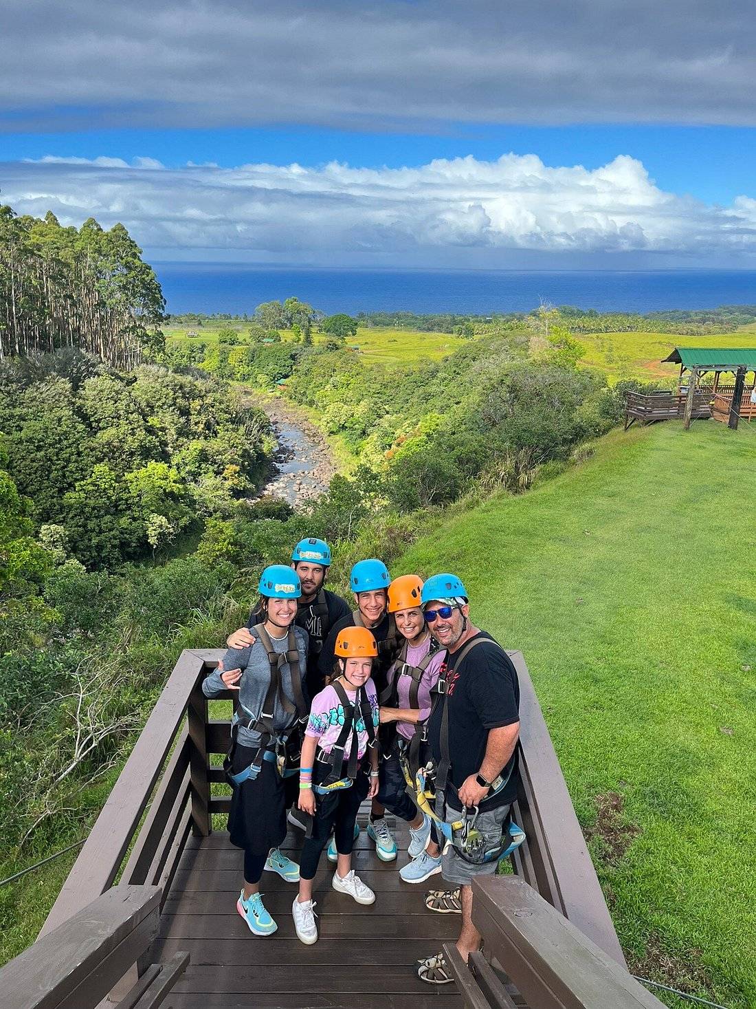 Family group in helmets and harnesses posing on a zipline platform overlooking the river valley and ocean at Umauma.