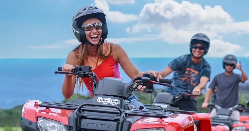 Three helmeted riders drive red ATVs along a dirt trail with lush green hills and the blue Pacific Ocean visible behind them on Hawaii Island.