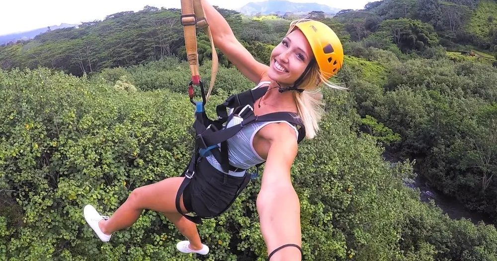 Smiling helmeted zipline rider glides above dense green Kauaʻi forest with mountain ridges rising in the background.