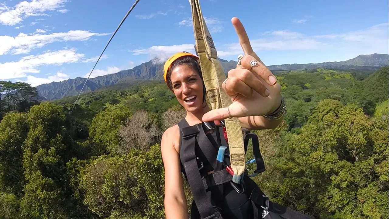 Guest in a zipline harness throwing a shaka with Kauaʻi's mountain ridgeline and jungle valley behind her