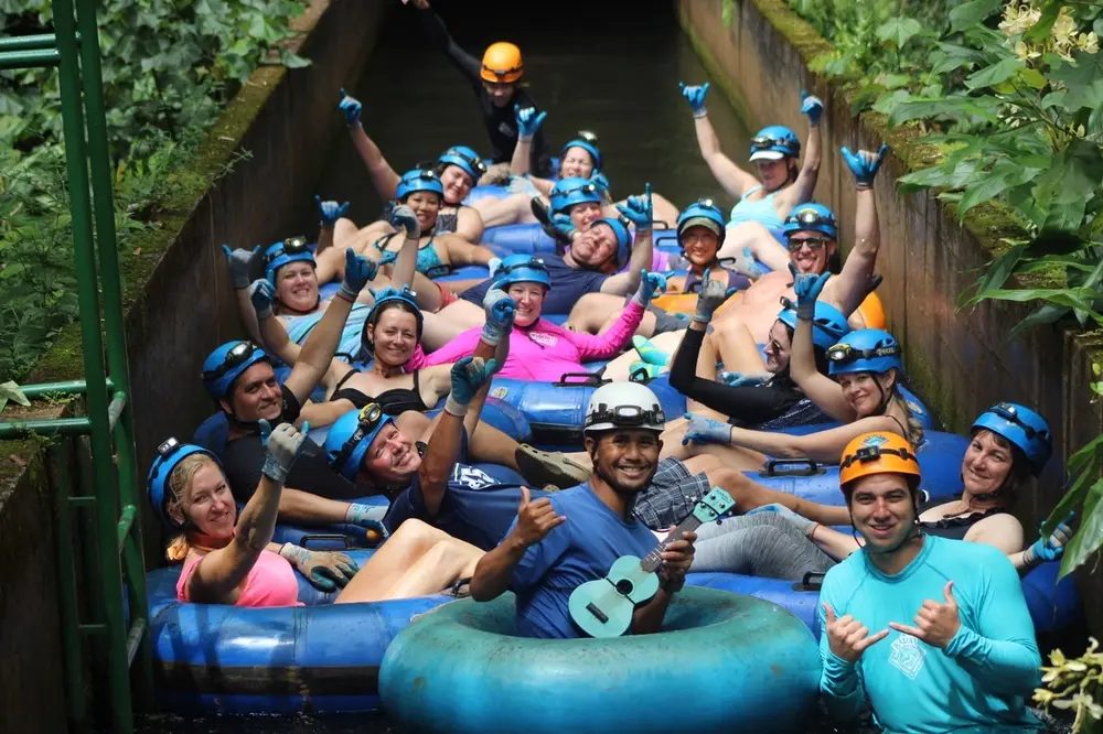 Large group of guests in helmets and gloves floating on inner tubes in a row, cheering as they approach one of the historic hand-dug tunnels on the Kauaʻi mountain tubing course.