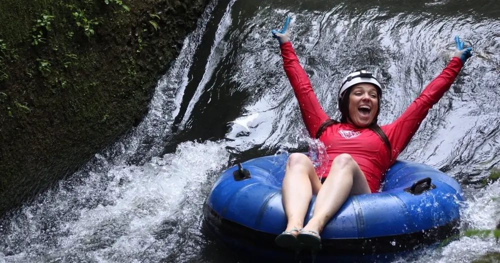 Guest in a red rash guard and helmet throws both arms up while riding an inner tube down a small waterfall drop in one of the plantation canals.