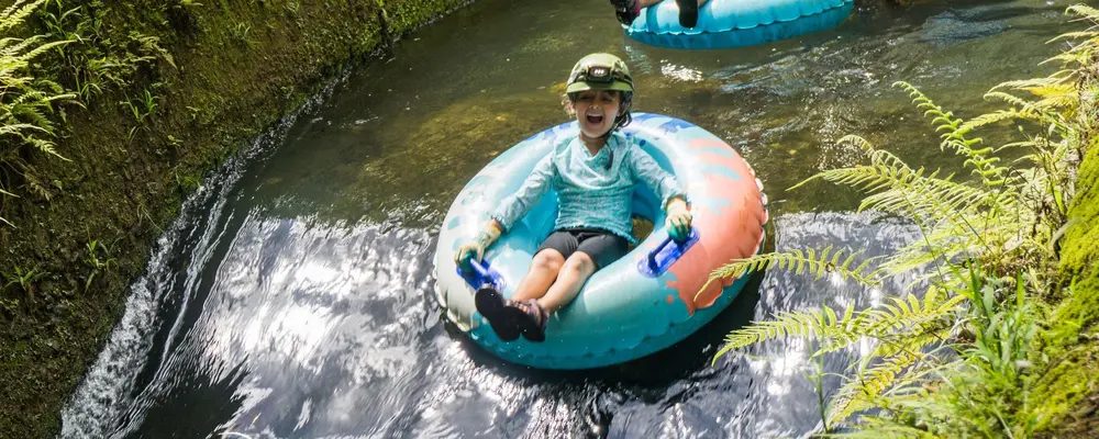 Young child laughing while floating alone in an inner tube through a calm, fern-lined jungle canal on the family-friendly Kauaʻi mountain tubing tour.