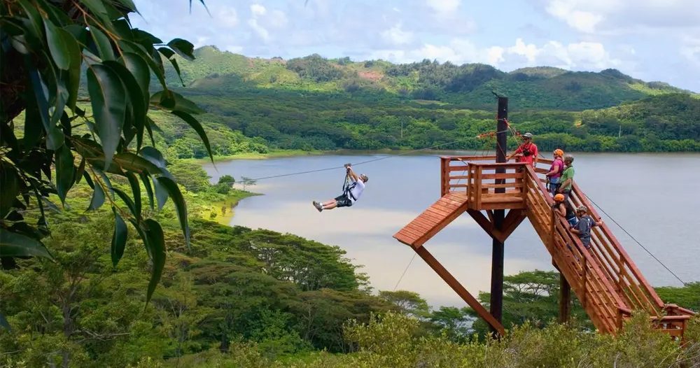 Zipline rider glides away from a wooden launch tower above the green shoreline of Waita Reservoir, with forested Kauaʻi hills rising in the distance.