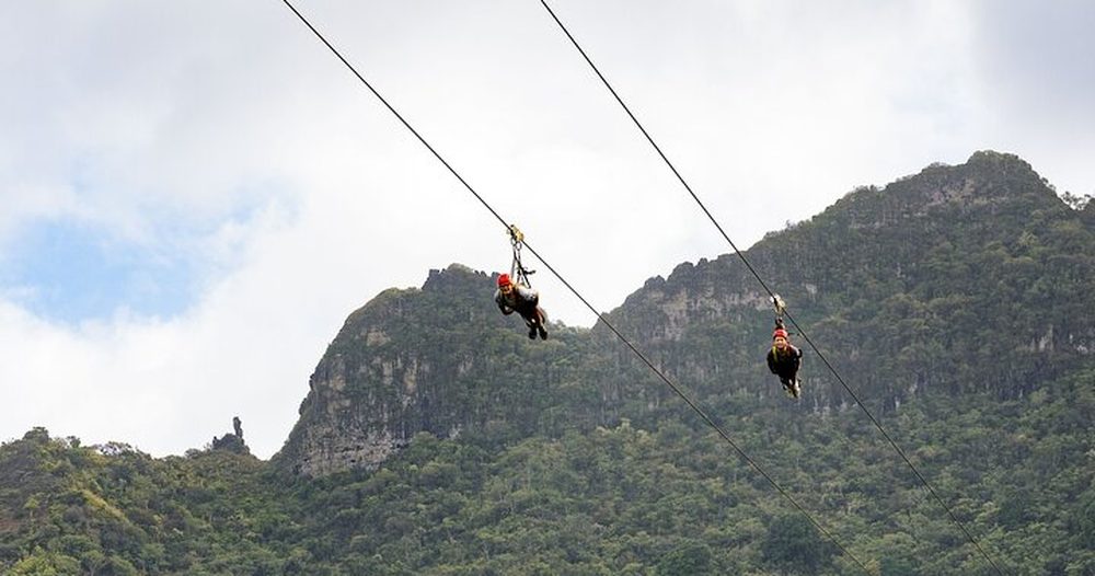 Two zipliners racing side by side on the tandem line, suspended above a lush green Kauaʻi valley with mountain peaks in the background.