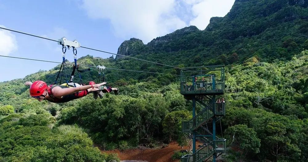 Two helmeted riders fly side by side in Superman-style harnesses above dense green forest, with a launch tower and steep Kauaʻi mountain ridges behind them.