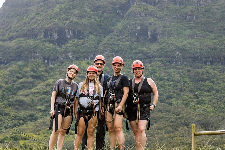 Group of five guests in helmets and zipline harnesses posing together in front of a forested Kauaʻi mountainside before their tour.