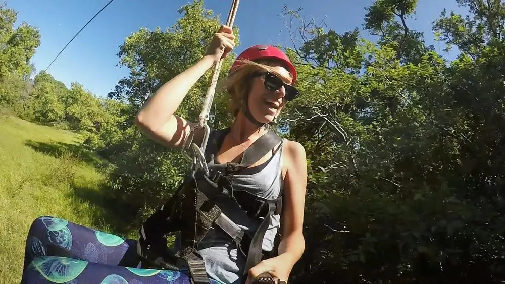 Smiling guest in a red helmet and harness on a zipline platform, sunlit trees and blue sky in the background.
