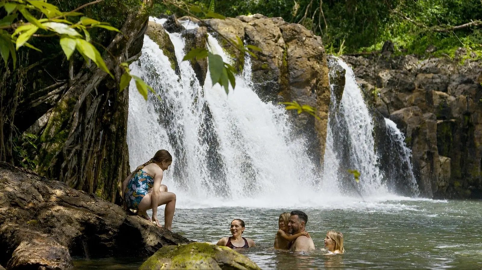 A family swimming and relaxing in the natural pool at the base of Kīpū Falls, with the waterfall cascading behind them.