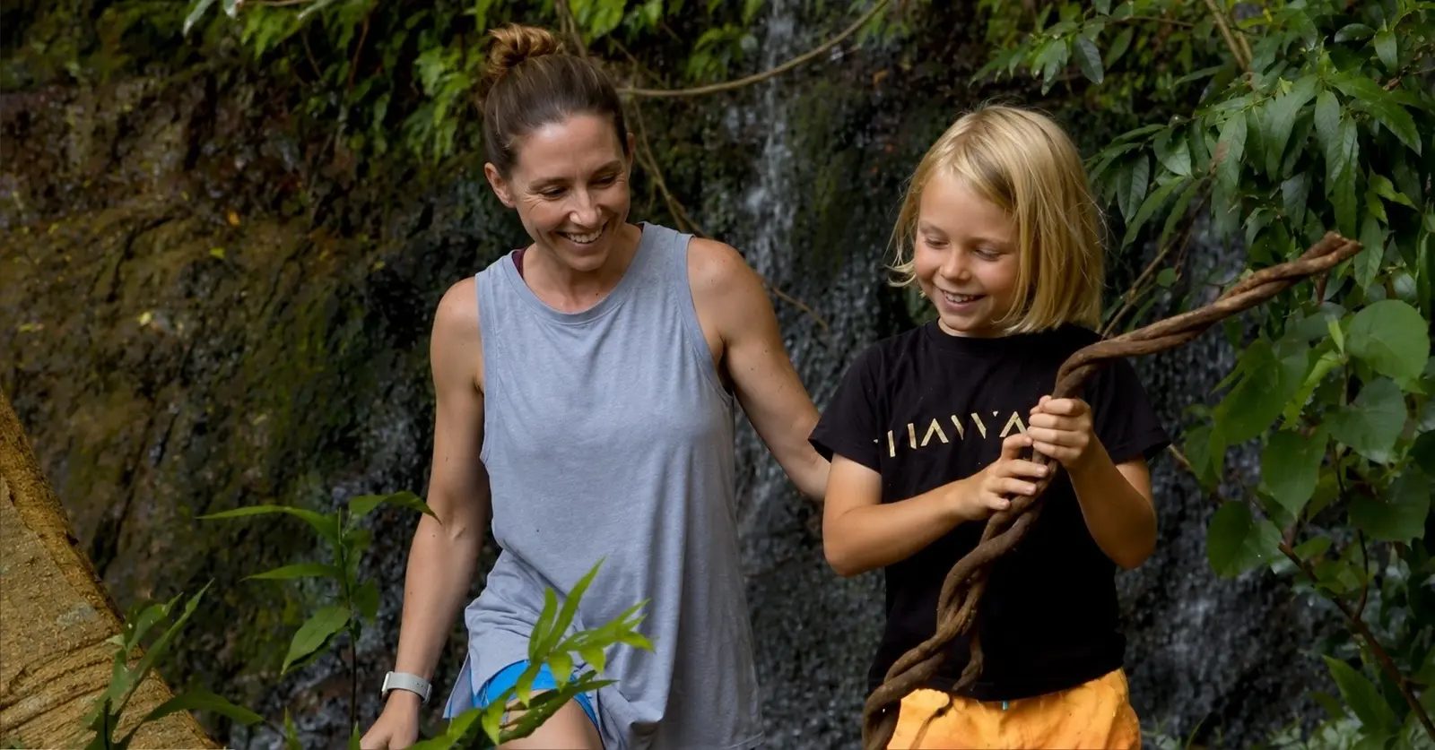 A guide and young hiker smiling and holding onto a jungle vine beside a waterfall along the Kīpū Falls trail.