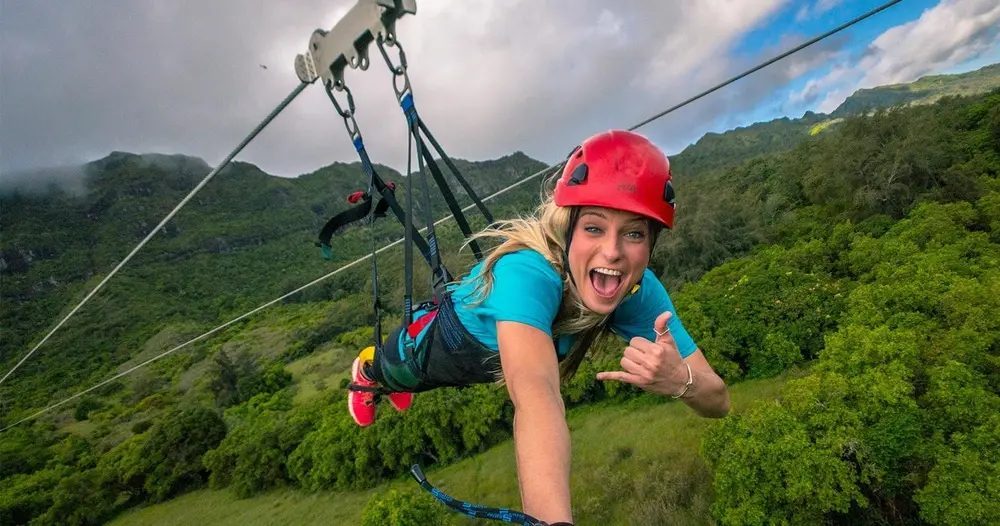 Smiling helmeted zipline rider flies Superman-style above lush green Kipu Ranch with steep Kauaʻi mountain ridges and low clouds in the background.