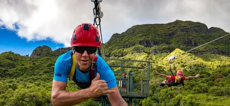 Guide giving a shaka while zip-lining toward the camera with a launch platform and another rider soaring across the Haʻupu Mountain Range behind him.