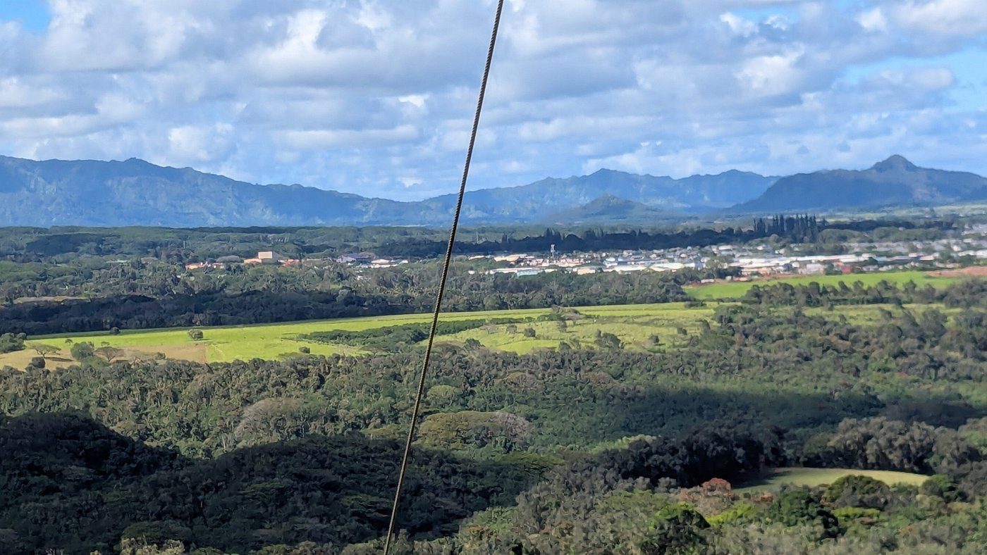Zipline cable stretching over Kipu Ranch's forested valley toward the Lihue plain and distant mountains.