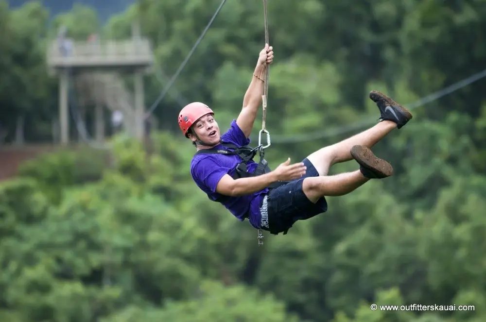 Guest swinging into a zipline platform with legs kicked up, jungle canopy blurred in the background.