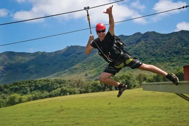 Zip-liner cheering with arms raised mid-flight over open pasture with Kipu Ranch's mountains in the background.