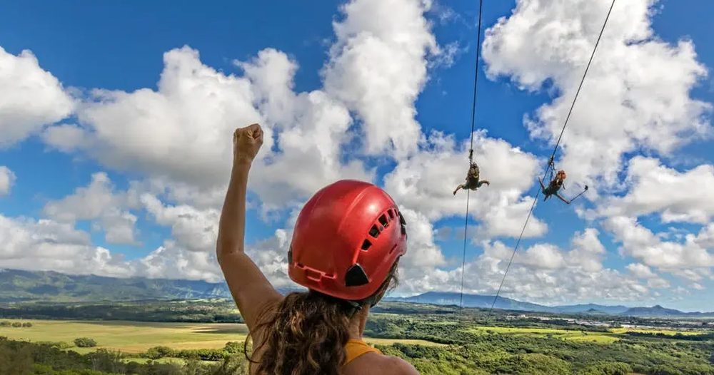 Helmeted guest raises a fist while two riders soar on parallel ziplines above the green fields and forested hills of Kipu Ranch under a bright Kauaʻi sky.