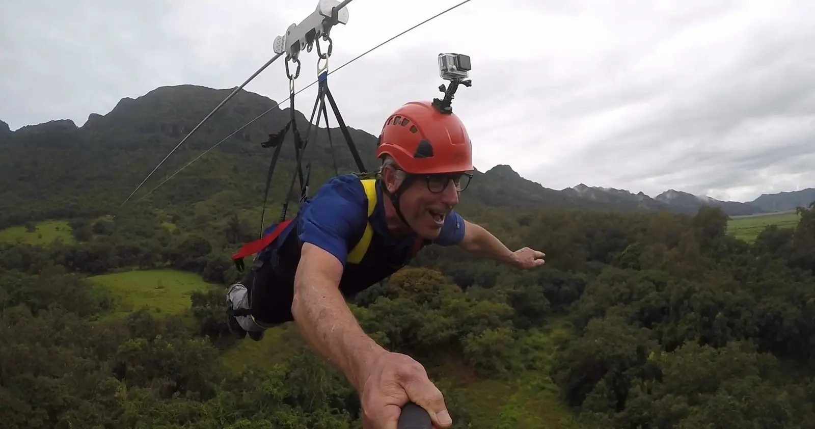 Flyer grinning while flying headfirst Superman-style on the FlyLine zipline, mountains and green valley below.