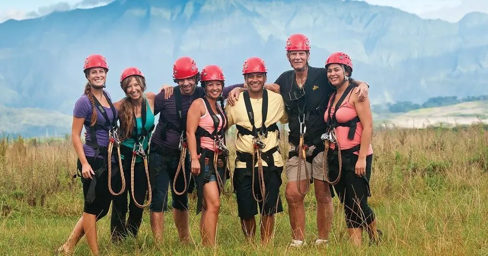 Seven helmeted guests wearing zipline harnesses stand together in a grassy field with the dramatic green Haʻupu Mountain Range rising behind them on Kauaʻi.