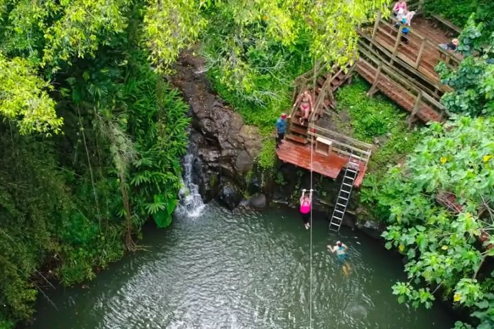 Aerial view of a guest riding Kauaʻi's only water zipline down into a jungle waterfall pool while another guest swims below.