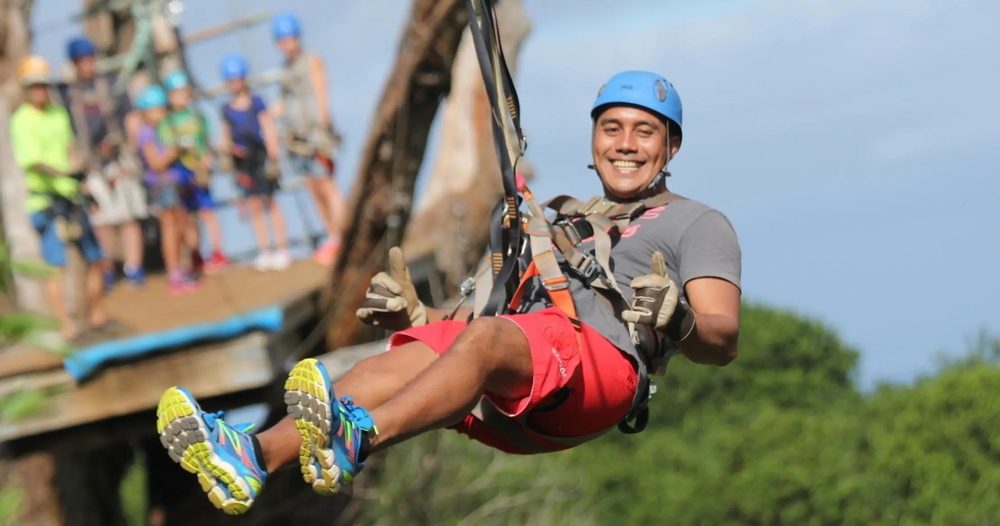 Guide smiling and giving a thumbs up while riding a zipline through the treetops on the NorthShore Zipline Tour, Maui