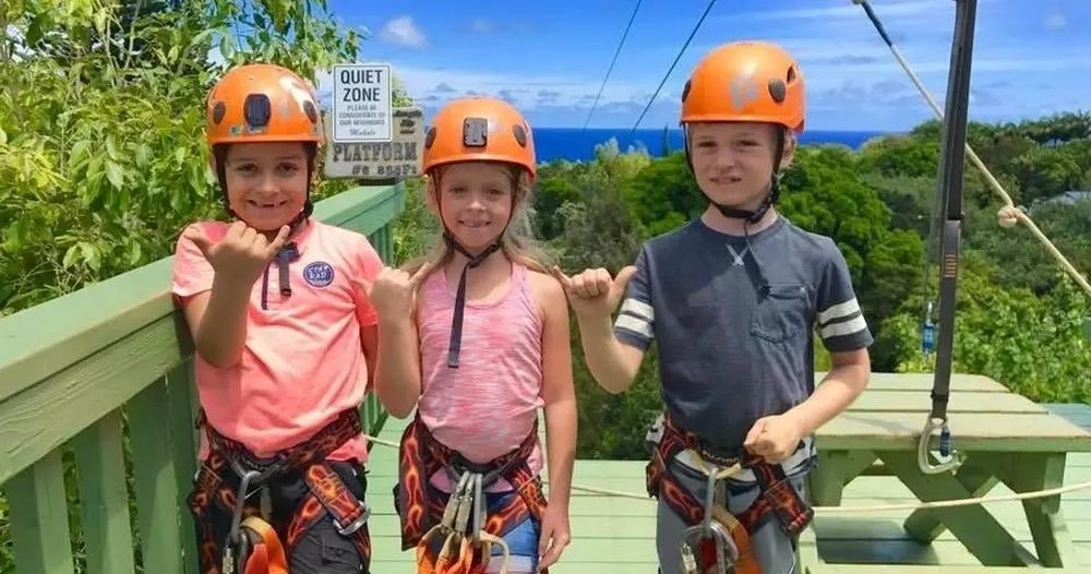 Three young zipline participants wearing orange helmets and harnesses stand on a raised platform with lush green forest and blue ocean visible behind them.