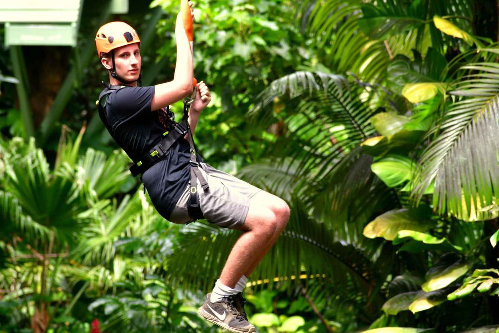 Zipline rider in an orange helmet gripping the trolley while gliding through the green jungle canopy on the 5 Line Zipline Eco Tour.