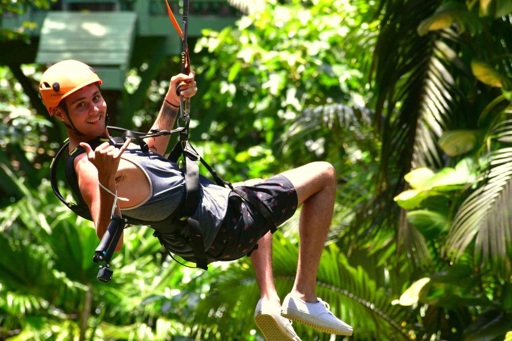 Smiling zipline rider giving a shaka sign while soaring through the rainforest canopy, surrounded by tropical foliage.