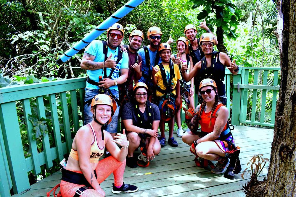 Group of guests in orange helmets and harnesses posing together on a jungle zipline platform, giving shaka signs.