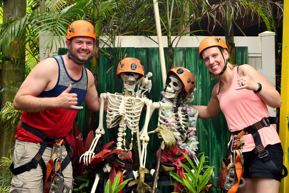 Two guests in zipline harnesses posing with the tour's costumed skeleton decorations near the check-in area.