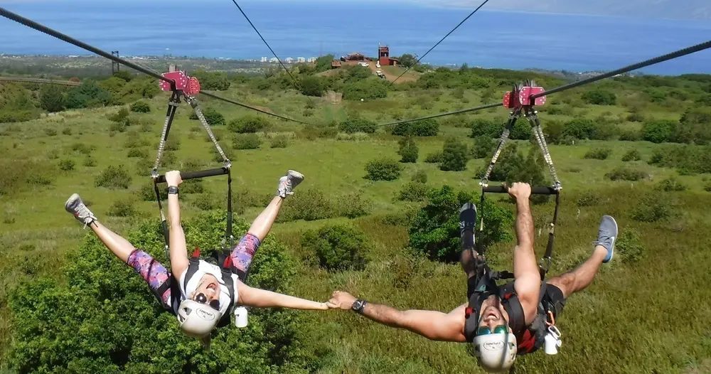 Two zipline riders hang upside down and hold hands while gliding side by side above a green Maui landscape with the blue ocean in the distance.